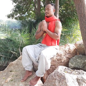 May include: A man wearing a red scarf sits cross-legged on rocks with his eyes closed and hands in a prayer position.