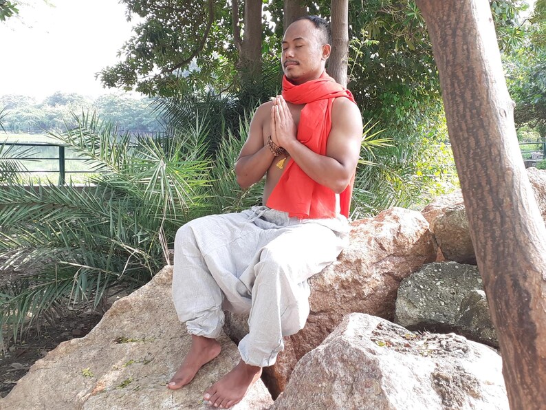 May include: A man wearing a red scarf sits cross-legged on rocks with his eyes closed and hands in a prayer position.