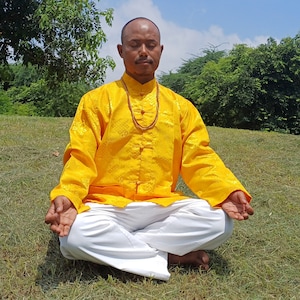 以下が含まれることがあります： 黄色の模様のある長袖シャツと白いズボンを着た男性が、草の生えた丘に座って目を閉じている。彼は、手をムドラのポーズにして瞑想している。