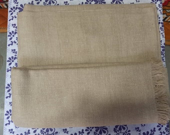 May include: A beige shawl with a fringe on one end. The shawl is folded in half and is lying on a white surface with a floral pattern.