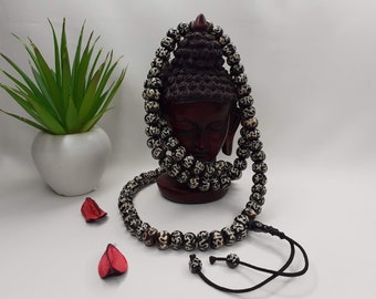 May include: A black and white prayer bead necklace with a decorative knot at the end. The beads are made of bone and have intricate carvings. The necklace is draped over a wooden Buddha statue.