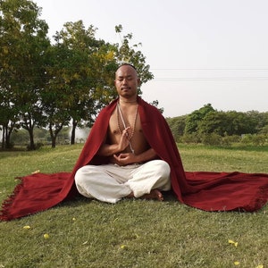 Pode incluir: Um homem meditando em um parque, vestindo um xale marrom e calças brancas. Ele está sentado de pernas cruzadas, com os olhos fechados e as mãos em posição de oração. Ele está usando um colar de contas.
