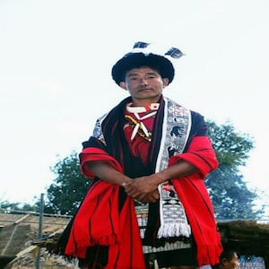 May include: A man wearing a traditional black and red patterned outfit with a white and black patterned sash. He is wearing a black hat with a white feather and has a serious expression on his face.