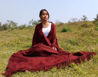 May include: A woman sits on a grassy hill wearing a maroon shawl with fringe. The shawl is draped around her shoulders and she is sitting with her legs crossed.