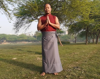May include: A man wearing a maroon sleeveless shirt with a grey long skirt and a beaded necklace. He is standing in a park with green grass and trees in the background. He is holding his hands together in a prayer position.