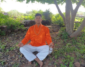 May include: A bright orange long-sleeved shirt with a mandarin collar and a subtle tonal pattern, paired with plain white trousers. The shirt features a front button closure and is worn with a long beaded necklace and a handheld string of prayer beads.