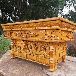 May include: A rectangular wooden altar table with a glossy honey-colored finish. The piece features intricate hand-carved floral patterns along the top edge and detailed relief carvings of dragons winding through clouds on the base and legs.