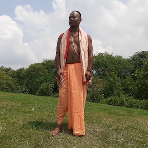 May include: A man wearing a traditional orange and white dhoti with a red and white scarf draped around his shoulders. He is standing in a grassy field with trees in the background.