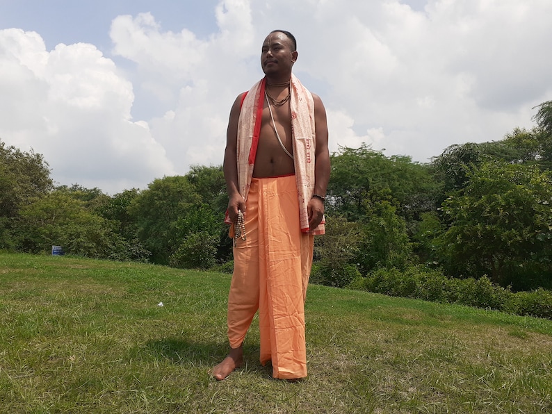 May include: A man wearing a traditional orange and white dhoti with a red and white scarf draped around his shoulders. He is standing in a grassy field with trees in the background.
