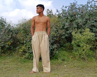 May include: A man wearing beige, loose-fitting pants with a drawstring waist. He is standing in a grassy field with bushes in the background.