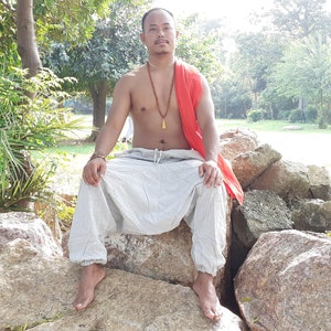 May include: A man wearing a white pair of loose-fitting trousers with a drawstring waist, a red scarf, and a gold necklace sits on a large rock. He is barefoot and has a relaxed expression on his face.
