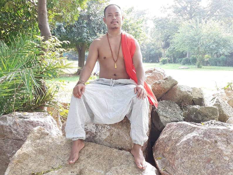 May include: A man wearing a white pair of loose-fitting trousers with a drawstring waist, a red scarf, and a gold necklace sits on a large rock. He is barefoot and has a relaxed expression on his face.