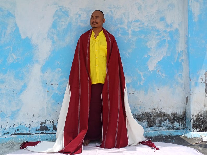 May include: A person wearing a yellow shirt and burgundy pants, draped with a red and white shawl. The shawl has vertical stripes and a fringed edge. The background is a blue wall with white accents.