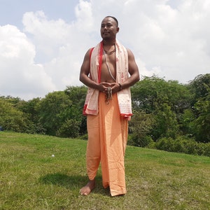May include: A man wearing a white and orange dhoti with a red and white patterned shawl draped over his shoulders. He is standing in a grassy field with trees in the background.