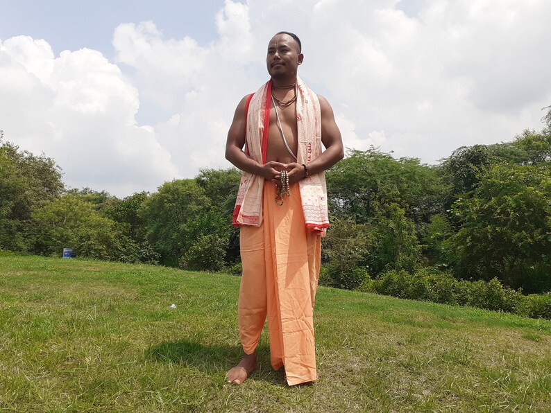 May include: A man wearing a white and orange dhoti with a red and white patterned shawl draped over his shoulders. He is standing in a grassy field with trees in the background.