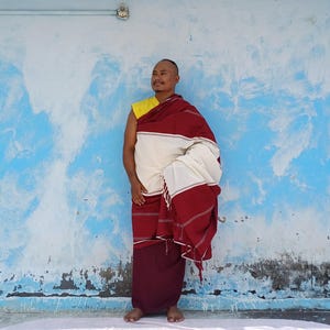 May include: A person wearing traditional Buddhist robes. The robes are a combination of maroon, white, and yellow. The person is standing in front of a blue wall with white paint streaks.