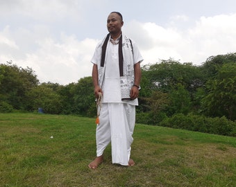 May include: A man wearing a white kurta and dhoti with a black and white patterned scarf. He is holding a beaded mala with an orange tassel.