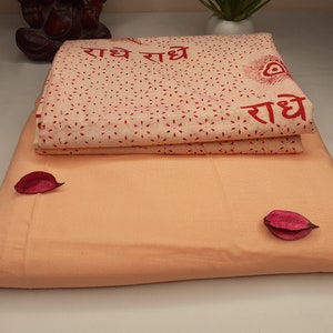 May include: A peach-colored cotton fabric with a white cotton fabric on top. The white fabric has a red print of the words "राधे राधे" and a peacock feather design.