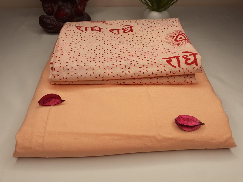 May include: A peach-colored cotton fabric with a white cotton fabric on top. The white fabric has a red print of the words "राधे राधे" and a peacock feather design.