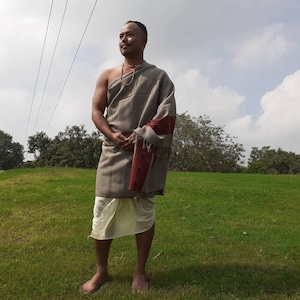 May include: A man wearing a gray shawl with red and white accents, standing in a grassy field. He is wearing a white underskirt and has a necklace around his neck.