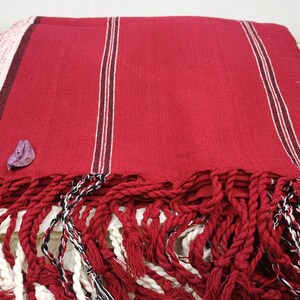 May include: A red woven textile with white and dark red stripes. The textile has a thick fringe in red, white, and black. A small purple petal rests on the fabric. This item is suitable for use as a blanket or throw.