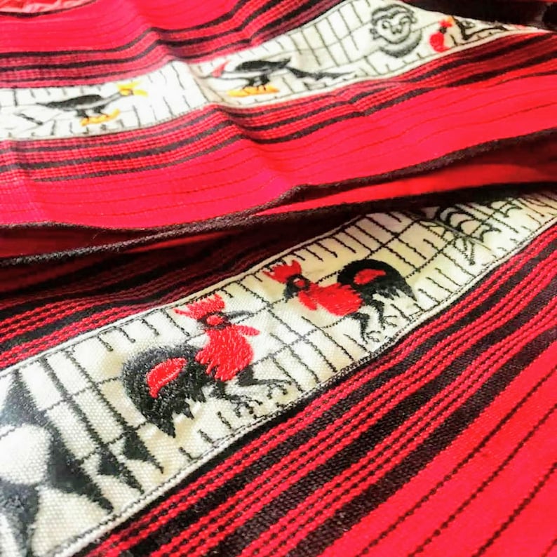 May include: Red and black striped fabric with white embroidered designs of roosters and other birds. The fabric has a woven texture.