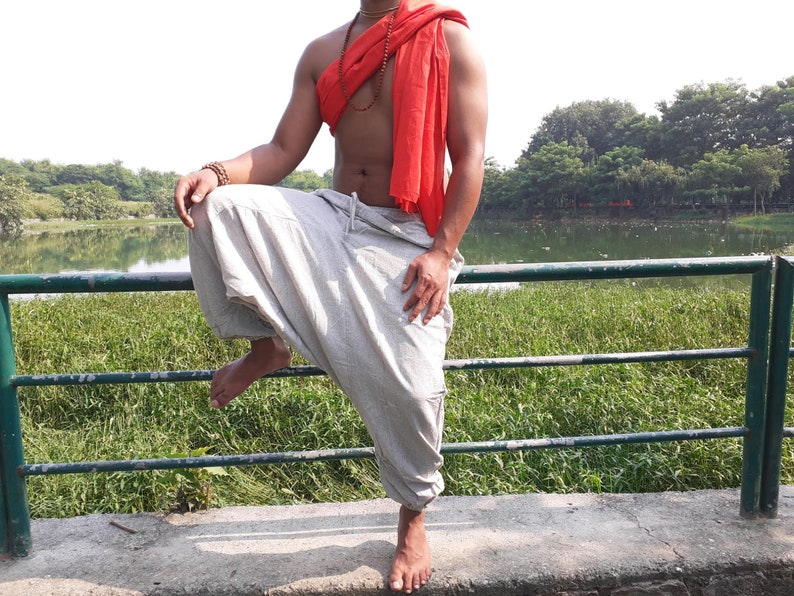 May include: A person wearing a white pair of loose-fitting trousers with a red scarf around their neck. The person is standing on a concrete surface with a green metal railing in front of them.