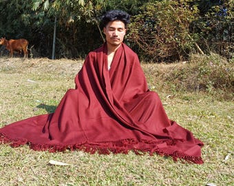 May include: A person sits cross-legged on the ground wearing a deep red shawl with fringe. The shawl is draped over their shoulders and covers their legs. The person is sitting in a grassy area with green foliage in the background.