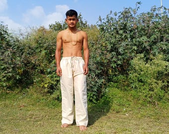 May include: A person wearing a pair of cream-colored pants with a drawstring waist. The person is standing in a field with green grass and bushes.