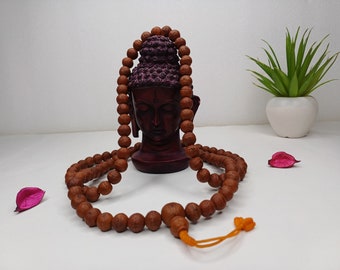 May include: A brown wooden mala bead necklace with a tassel, draped over a dark red Buddha statue with a purple crown. The necklace is on a white surface with pink flower petals.