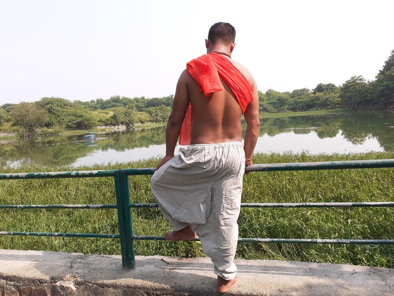 May include: A person wearing white loose-fitting trousers and a red scarf, standing by a railing overlooking a lake.