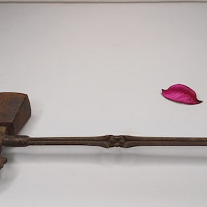 May include: A bronze hand-held ritual tool with two spinning wheels on each end and a square wooden handle. The tool is on a white surface with a pink flower petal.