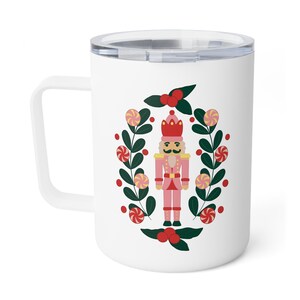 May include: White travel mug with a handle and a silver lid. The mug features a pink nutcracker illustration surrounded by a wreath of green leaves, red berries, and candies. A festive holiday design.