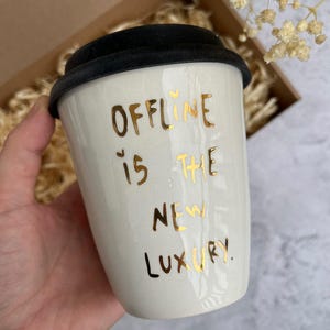 Puede incluir: Taza de viaje de cerámica blanca con tapa negra. La taza tiene letras doradas que dicen "Offline is the new luxury."