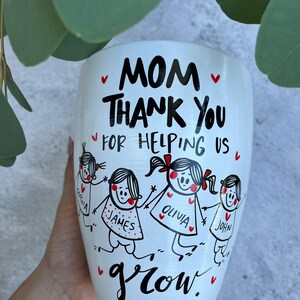 May include: White ceramic planter with a hand-drawn design. The planter features the text "MOM THANK YOU FOR HELPING US grow." Below the text are illustrations of children holding hands. Red hearts are scattered around the design.