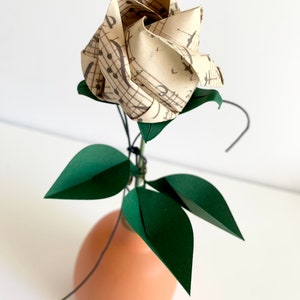 Angled side view of the Origami paper rose in a ceramic vase. The rose button made with a vintage looking musical sheets paper, shown from an angle that emphasizes  the three emerald dark green paper leaves, and the semi-opened twisted rose button.