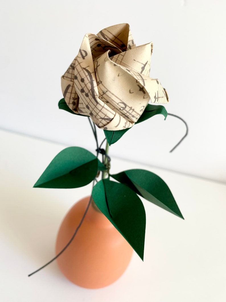 Angled side view of the Origami paper rose in a ceramic vase. The rose button made with a vintage looking musical sheets paper, shown from an angle that emphasizes  the three emerald dark green paper leaves, and the semi-opened twisted rose button.