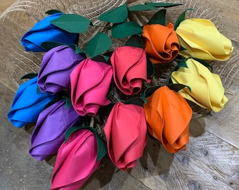 May include: A colorful floral arrangement featuring paper roses in shades of blue, purple, pink, orange, and yellow. The roses are arranged on a mesh base with green paper leaves, creating a vibrant and eye-catching display.