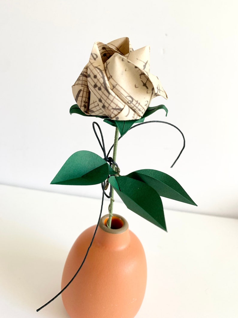 View of the Origami paper rose in a ceramic vase. The rose button made with a vintage looking musical sheets paper, shown from an angle that emphasizes  the three emerald dark green paper leaves, and the semi-opened twisted rose button.