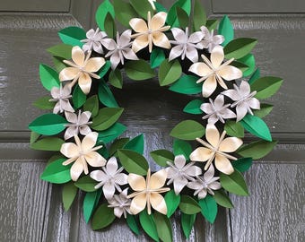May include: A handmade wreath featuring paper flowers and leaves. The wreath has a circular shape with green leaves and cream and silver-colored flowers. The wreath is hanging on a dark green door. The text "@BEEMELLOFLOWERS" is visible.