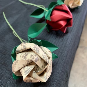 View of various origami roses laying on a table against a black cloth to emphasize its vintage looking color. The first rose folded with the Musical sheets paper, the second one is a red rose  and the third one made with the I love you printed paper.