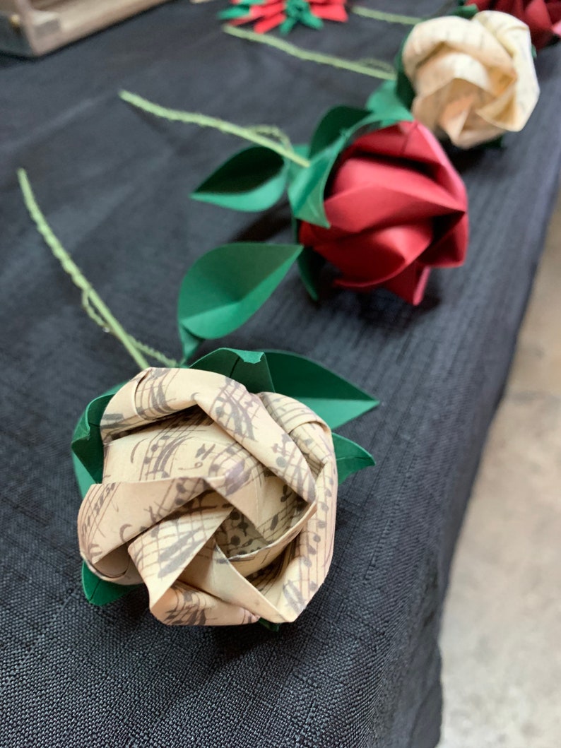 View of various origami roses laying on a table against a black cloth to emphasize its vintage looking color. The first rose folded with the Musical sheets paper, the second one is a red rose  and the third one made with the I love you printed paper.