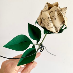 View of the Origami paper rose in my hand.  Showing the rose button vintage looking musical sheets paper and the three emerald dark green paper leaves. You can also view the small metal heart added to the stem with a small string.