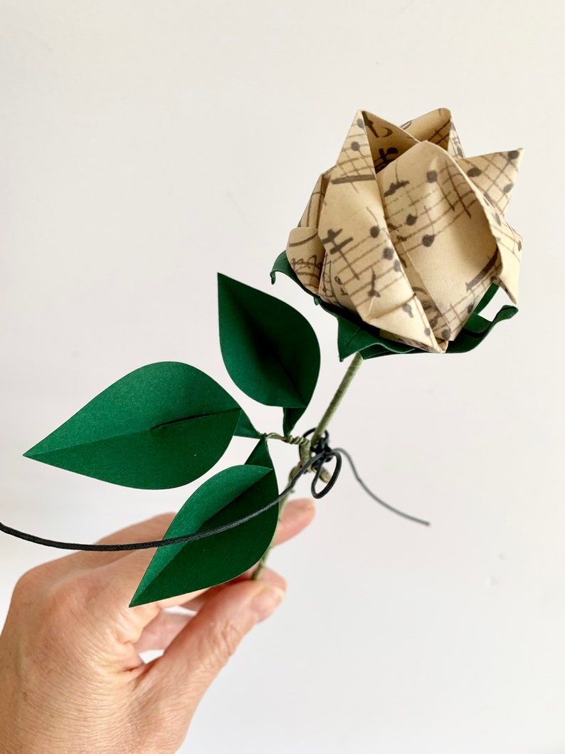 View of the Origami paper rose in my hand.  Showing the rose button vintage looking musical sheets paper and the three emerald dark green paper leaves. You can also view the small metal heart added to the stem with a small string.