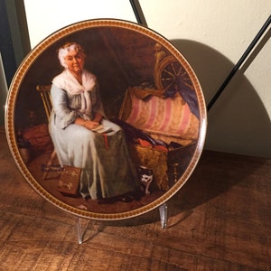 May include: A decorative porcelain plate featuring a painting of a woman sitting in a chair with a cat at her feet. The plate has a gold rim and a brown background. The text "Norman Rockwell" is printed on the bottom of the plate.