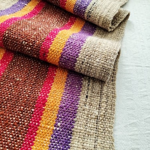 Peut inclure: Un chemin de table tissé avec un motif rayé dans des tons de brun, orange, rose et violet. Le chemin de table est fait d'une fibre naturelle et a un aspect rustique et tissé à la main.