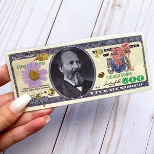 May include: A $500 bill with a portrait of a man with a beard and a flower on the left side. The bill has the text "THE UNITED STATES OF AMERICA" and "FIVE HUNDRED" on it. The bill is in a clear plastic sleeve.