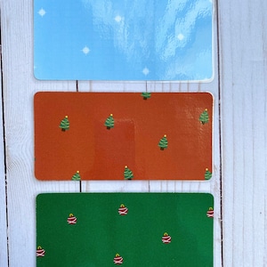 May include: Three rectangular stickers with different holiday designs. The top sticker is light blue with white snowflakes. The middle sticker is orange with a repeating pattern of green Christmas trees. The bottom sticker is green with a repeating pattern of red ornaments.