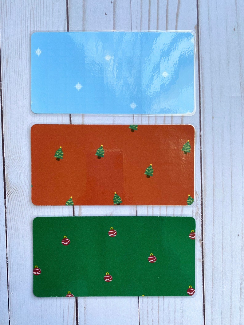 May include: Three rectangular stickers with different holiday designs. The top sticker is light blue with white snowflakes. The middle sticker is orange with a repeating pattern of green Christmas trees. The bottom sticker is green with a repeating pattern of red ornaments.