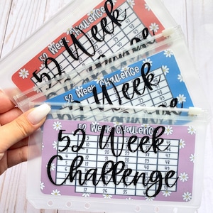 May include: Three clear plastic zippered pouches with a 52 week challenge tracker inside. The trackers are printed on pink, blue, and red backgrounds with white daisies. The text on the trackers reads "52 Week Challenge".
